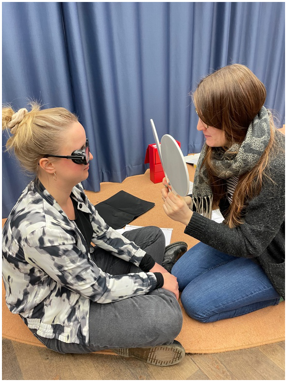 Das Foto zeigt eine Überprüfung des Sehvermögens („Preferential Looking“). Zwei Frauen sitzen sich auf einem Teppich in Armlänge gegenüber. Eine Frau trägt eine Simulationsbrille und schaut auf zwei Paddles, die die andere Frau auf Augenhöhe präsentiert.