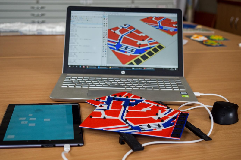 The picture shows a laptop demonstrating the modelling of a tactile map and the construction of a mobile application. A tablet is connected to the laptop and two interactive tactile maps are placed around it.