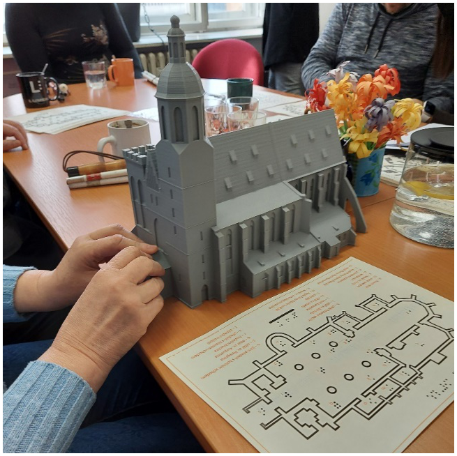 The picture is a photo taken during the testing of the 3D models. The photo shows a table on which there is a 3D model of the cathedral and its floor plan, which is printed on a thermal printer. The model of the cathedral is being felt by two hands of a research participant who took part in the testing of the tools.