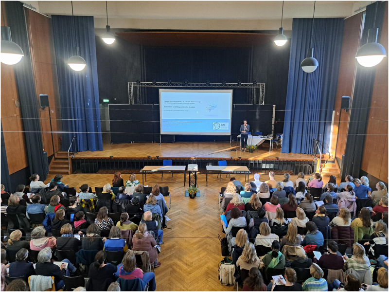 Blick von oben: Die Aula der LVR-Louis-Braille-Schule ist gut gefüllt. Frau Dr. Unterberger steht auf der Bühne, rechts neben der Leinwand.