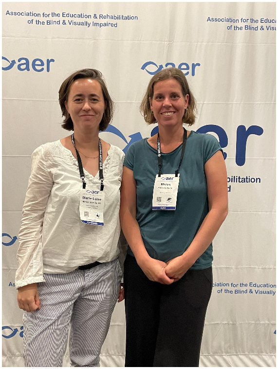 Das Foto zeigt Dr. Marie-Luise Schütt (links) und Maren Balschat (rechts) mit ihrem Konferenz-Badge um den Hals. Sie stehen vor dem Banner der AER-Konferenz und lächeln in die Kamera.