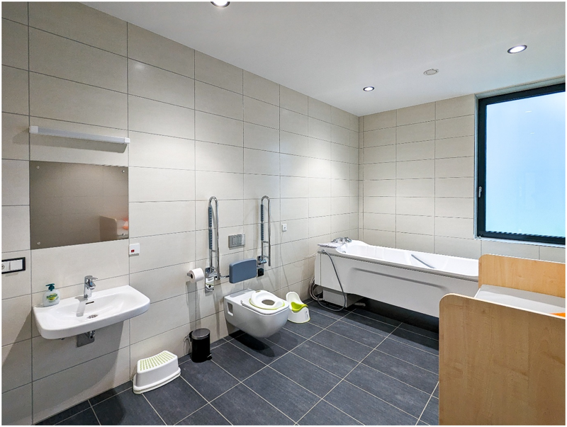 Großes barrierefreies Badezimmer mit Badewanne, Toilette mit Haltegriffen, Waschbecken, Wickeltisch, Fenster.