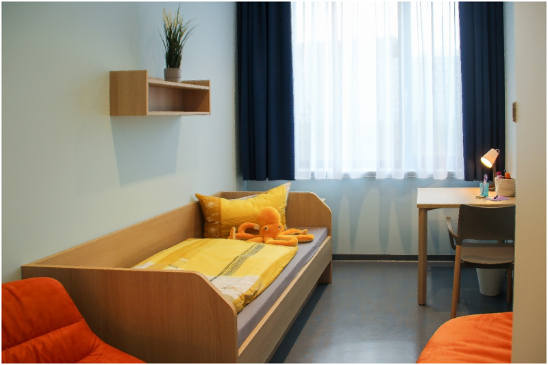 Einzelzimmer mit Bett, Kuscheltier, Schreibtisch, Regal, Sessel und Fenster mit Vorhang, Blick leicht von vorn.
