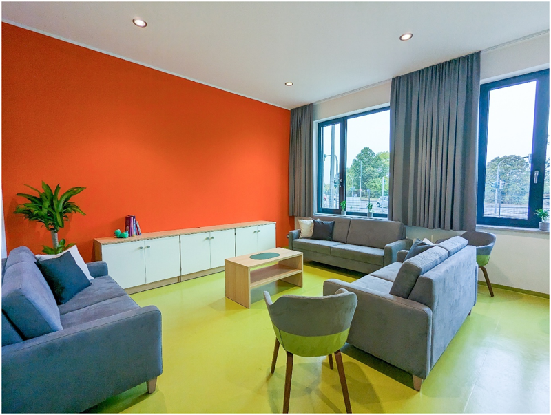 Aufenthaltsraum mit 4 Sofas, Sessel, kleinem Tisch, Sideboard, Pflanzen, Bücher und 2 Fenstern, frontal aufgenommen.