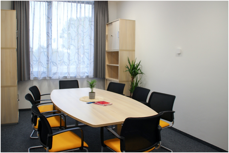 Teppich: Konferenzraum mit ovalem Tisch, 8 Stühlen, einem Schrank, Regal, Pflanze und Fenster mit Vorhang, Blick leicht schräg von vorne.