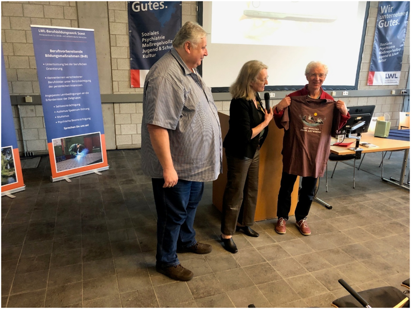 Frank Laemers, Kirsten Wahren-Krüger und Horst Hülsmann stehen nebeneinander in der Aula des LWL-Bildungszentrums Soest. Horst Hülsmann lächelt und hält sich ein braunes T-Shirt vor die Brust mit der Aufschrift „Not working from anywhere“ und dem Bild einer Person in einer Hängematte.