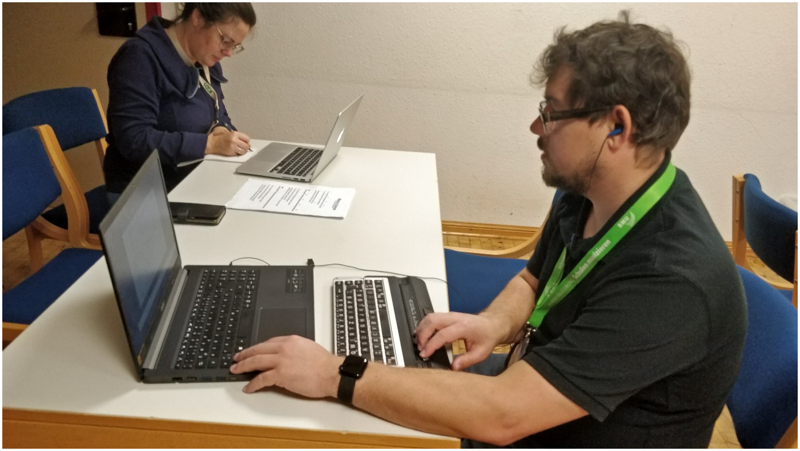 An einem Tisch im Seminarraum sitzen 2 Personen, die das Notenschreibprogramm MuseScore ausprobieren. Der Mann im Vordergrund nutzt zur Arbeit einen Laptop mit angeschlossener Braillezeile sowie Kopfhörer. Die Frau im Hintergrund arbeitet sowohl mit Laptop als auch mit Papier und Stift.