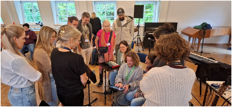 Eine Gruppe von Menschen steht und sitzt um einen jungen Mann herum, der in der Mitte des Raumes kniet und mit dem Finger auf einem iPad Bewegungen ausführt. Im Raum befinden sich u.a. mehrere Tasteninstrumente und Lautsprecher.