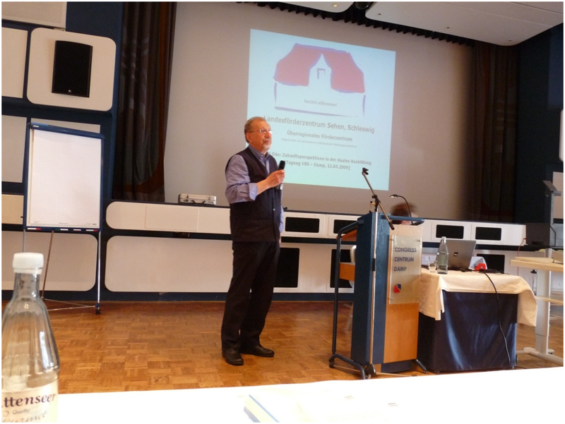 Das Foto zeigt Willi Düe als Sprecher bei einer Tagung der Einrichtungsleitungen aus dem Jahr 2009 in Damp. Im Hintergrund ist die Leinwand mit einem Beitrag sowie dem projizierten Logo vom LFS deutlich zu erkennen.