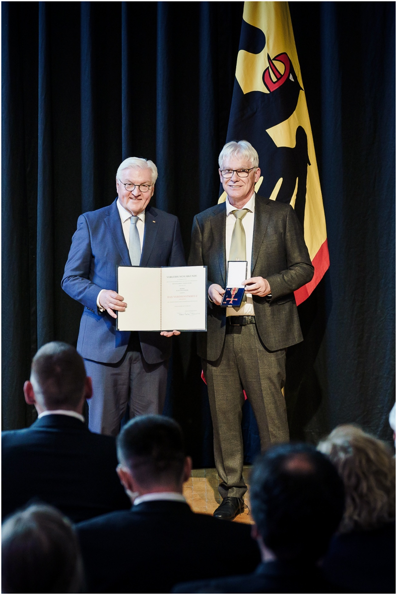 Bundespräsident Frank-Walter Steinmeier und Claus Duncker stehen auf der Bühne in der Stadthalle in Stadtallendorf vor einem dunkelblauen Vorhang und einer Deutschlandflagge. Bundespräsident Steinmeier hält die Mappe mit der Urkunde in den Händen. Claus Duncker steht rechts von ihm und hält die Schatulle mit dem Bundesverdienstkreuz. Beide tragen Anzug und Krawatte. Das Publikum im Bildvordergrund blickt zur Bühne.