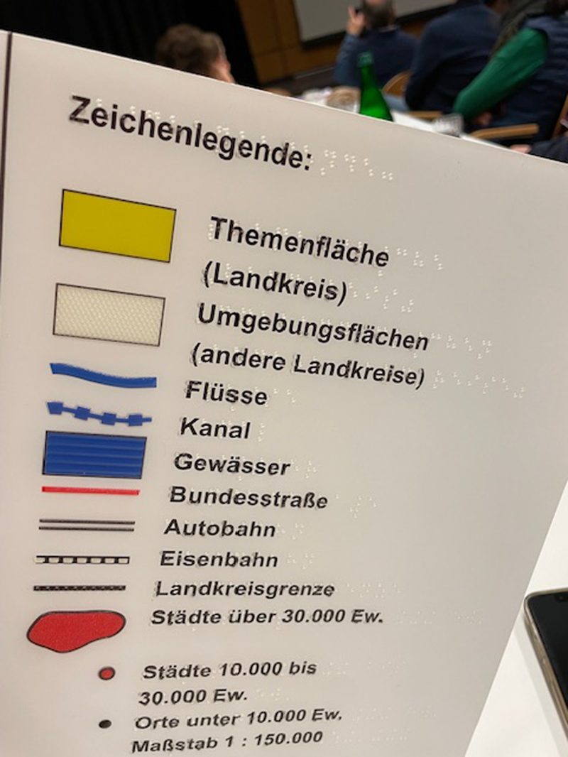Zeichenlegende einer taktilen Karte – auf der linken Seite der Legende sind taktile Auszüge für Flächen, Flüsse, Bundesstraßen, Autobahnen etc., die auch eingefärbt sind. Rechts daneben stehen die Schwarzschriftbeschreibungen, auf denen farblos die Punktschriftbeschriftungen zu sehen sind.