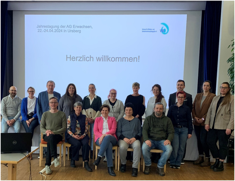 Die Teilnehmerinnen und Teilnehmer haben sich zu einem Gruppenbild aufgestellt. In der ersten Reihe sitzen fünf von ihnen auf Stühlen, die anderen stehen dahinter. Auf einer großen Leinwand im Hintergrund steht links oben „Jahrestagung der AG Erwachsen, 22.-24.04.2024 in Ursberg, rechts oben befindet sich das Logo des VBS. In der Mitte der Leinwand steht „Herzlich willkommen“.