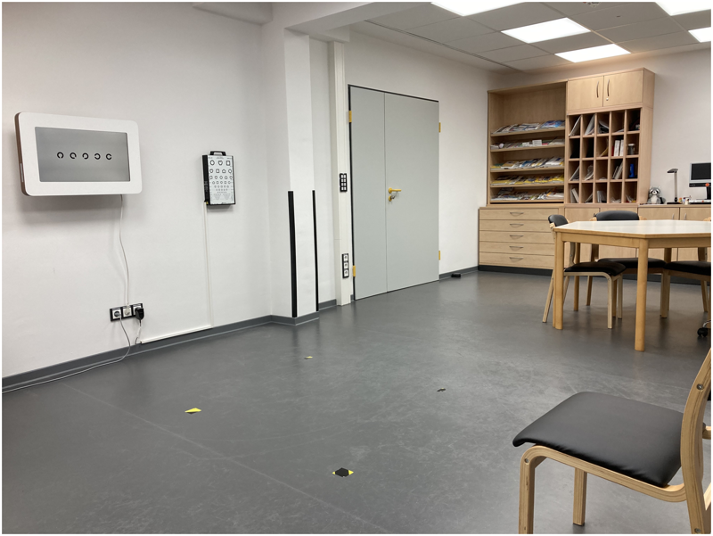 Teile des Diagnostikraums mit an der Wand befestigten Prüfgeräten zur Visusermittlung und einem in 3 Meter Testentfernung positionierten Stuhl sind auf dieser Abbildung dargestellt. Im Hintergrund befinden sich eine Tischgruppe mit einzelnen Stühlen sowie Schränke mit weiteren Arbeits- und Infomaterialien.    