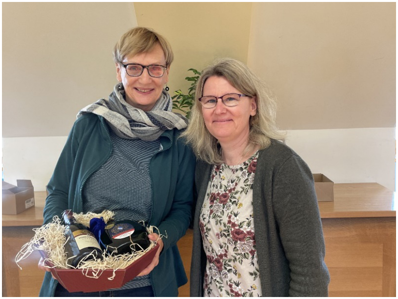 Abb.2 zeigt Mechthild Gahbler und Imke Wißmann. Die beiden stehen nebeneinander und lächeln in die Kamera. Mechthild Gahbler hält vor sich einen Geschenkkorb, gefüllt mit 2 Flaschen und einem blauen Säckchen. Im Hintergrund sieht man den Raum mit einer Dachschräge.
