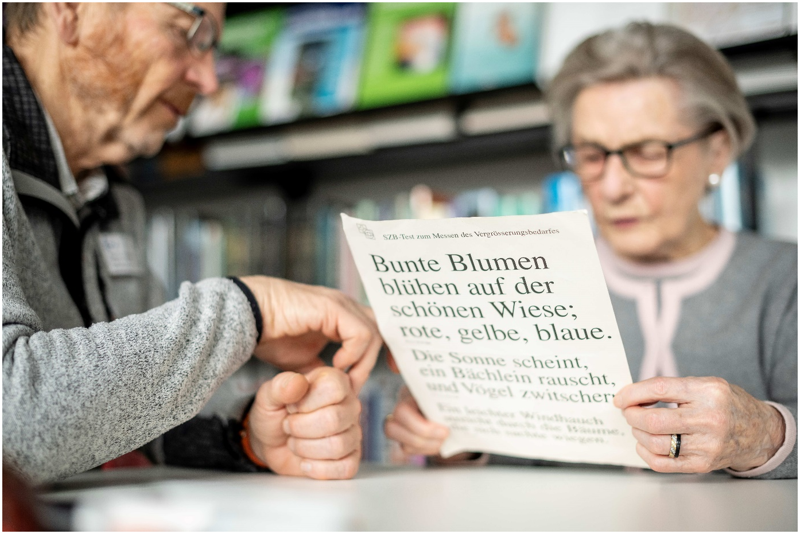 Foto 1: Ein Mann und eine Frau sitzen am Tisch. Die Frau liest von einem Blatt Papier ab, auf dem drei Sätze in unterschiedlichen Schriftgrößen, von groß nach klein, abgedruckt sind. Der Mann zeigt mit seiner Hand auf das Blatt.