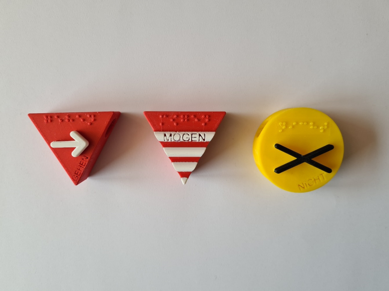 Foto; 3D-Symbole ‚gehen‘, ‚mögen‘, ‚nicht‘ liegen nebeneinander von links nach rechts. ‚gehen‘ enthält als taktiles Symbol einen weißen Pfeil, ‚mögen‘ vier weiße Rillen jeweils auf einem dreieckigen, roten Korpus. ‚nicht‘ enthält ein schwarzes x als taktiles Symbol auf einem runden, gelben Korpus.