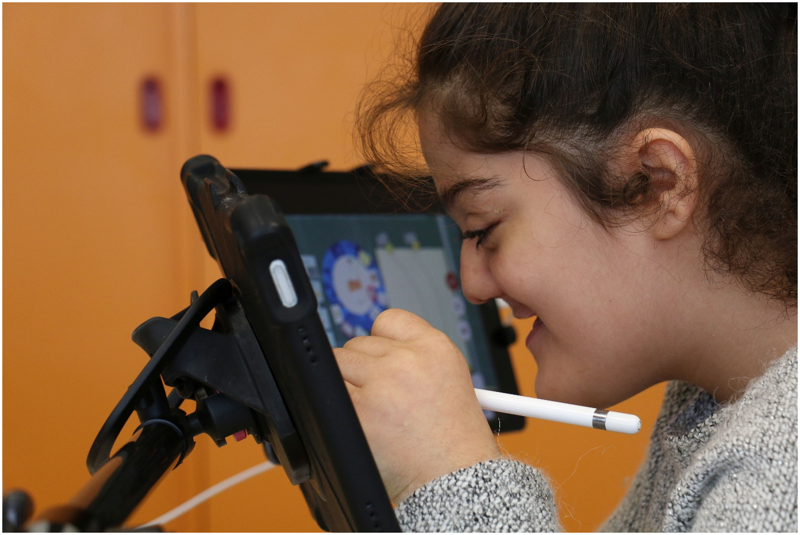 Auf dem Foto sieht man Serin im Profil vor zwei iPads auf einem Stativ, die im 110 Grad Winkel vor ihr angeordnet sind. Sie schreibt auf dem linken iPad mit dem Apple Pencil. Auf dem rechten iPad sieht man den Klassenraum aus dem Blickwinkel des Avatars. 
