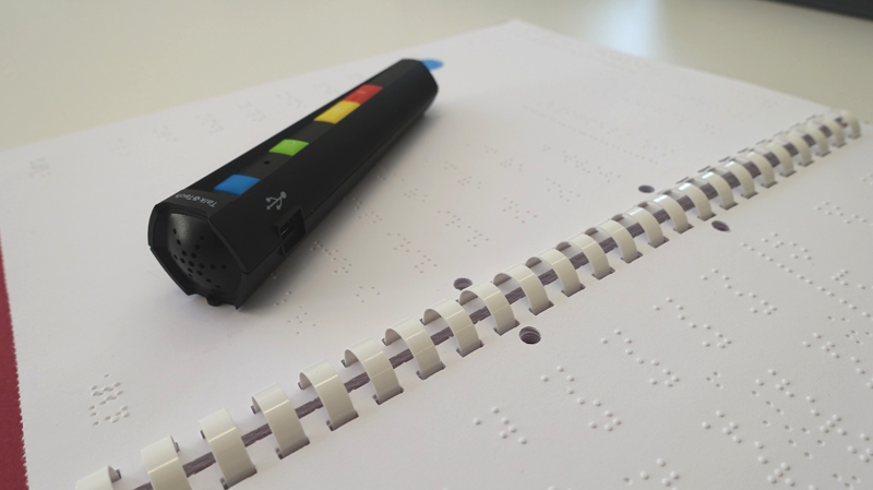 Portrait: Der PennyTalks-Hörstift liegt auf einer Seite der in Punktschrift übertragenen Fibel. Der etwa 16cm lange Stift läuft vom oberen, dickeren Ende (Durchmesser ca. 4cm) zur Spitze hin konisch zu. Am oberen Ende ist der eingebaute Lautsprecher zu erkennen. Auf dem Schaft des Stifts befinden sich eine blaue Anzeigeleuchte und drei Bedienelemente (grün, gelb und rot).