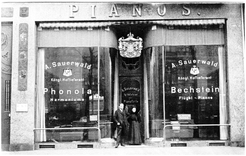 Historisches Foto in Sepiatönung von Anton Sauerwalds Musikalienhandlung aus dem Januar 1908. Im Vordergrund Anton Sauerwald, dahinter zwei Angestellte. 