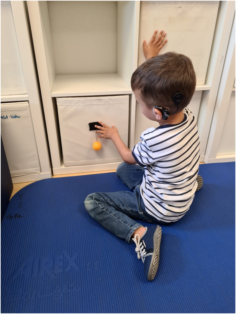 Das Foto zeigt einen Jungen mit CIs sitzend auf einer Matte vor seiner Spielzeugkiste von der Seite. Mit seiner linken Hand exploriert er das Schaffell, die rechte stützt er auf einer anderen Kiste im Regal ab.