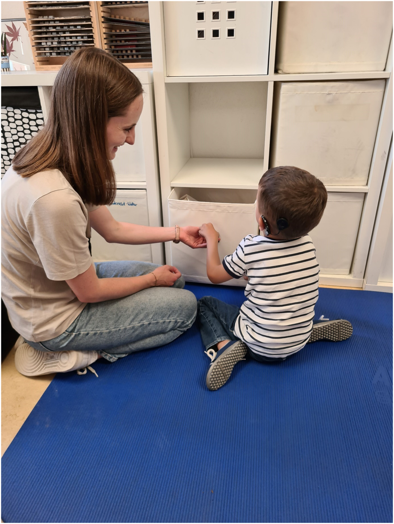 Auf dem Foto sitzt ein Junge mit CIs zusammen mit seiner Bezugsperson auf der Matte. Sie halten über die Knie Körperkontakt. Sie erkunden mit ihren linken Händen den Tischtennisball, der an seiner Spielzeugkiste zum leichteren Herausziehen befestigt ist. Die Spielzeugkiste ist bereits ein kleines Stück aus dem Regal gezogen.