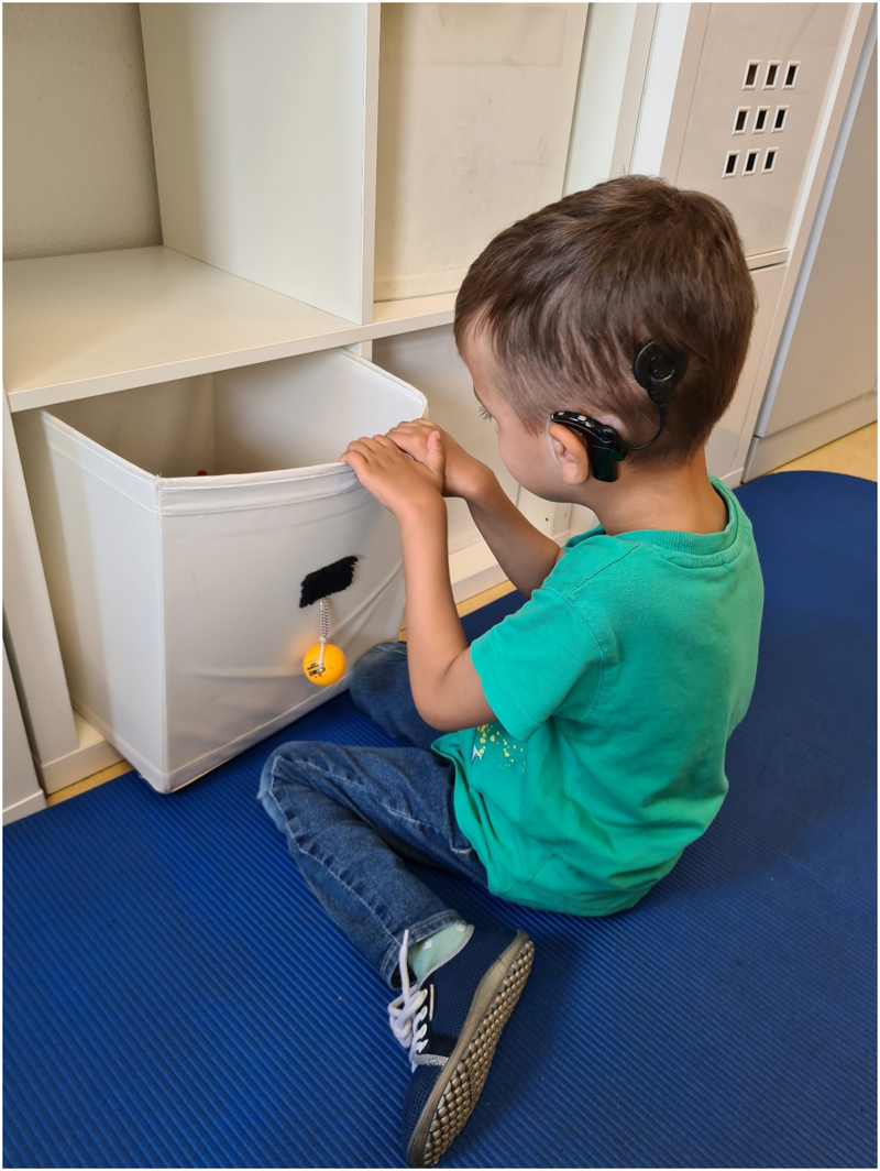 Auf dem Foto zieht der Junge mit CIs auf seiner Matte sitzend seine Spielzeugkiste aus dem Regal heraus, indem er mit beiden Händen an der oberen Kante der Kiste zieht.