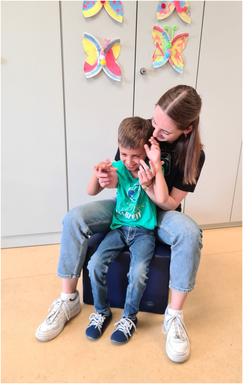 Auf dem Foto sitzt die Bezugsperson in engem Körperkontakt hinter dem Jungen mit CI auf einem Schaumstoffwürfel. Die Bezugsperson trägt eine digitale Übertragungsanlage. Sie sind in Interaktion, ihre Hände berühren sich, beide lachen.