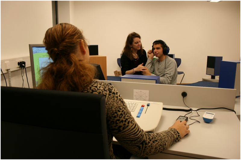 Foto: Im Vordergrund sitzt eine Frau am Computer. Sie schaut zu einem Jugendlichen mit Kopfhörern auf dem Kopf. Neben ihm sitzt eine Frau, die ihn anschaut und deren Hand der Jugendliche hält. Mit seiner anderen Hand gebärdet der Jugendliche „hören“. Der Jugendliche wirkt konzentriert.