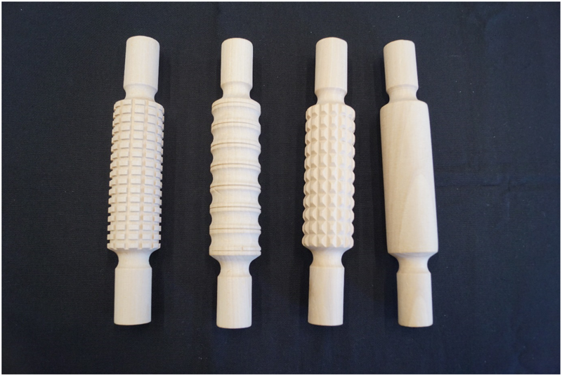 Auf einem schwarzen Stofftuch liegen vier, circa 25cm lange Holzwalzen. Im Mittelteil jeder Walze befindet sich ein spezifisches Relief (von rechts nach links): Eine Holzwalze ohne Relief, eine Holzwalze mit vielen kleinen konisch zulaufenden Stellen mit Rillen dazwischen, eine Holzwalze mit acht geraden Quererhebungen und Quervertiefungen und eine Holzwalze, mit vielen kleinen rechteckig-erhobenen Stellen mit Rillen dazwischen. Links und rechts der Reliefs weist das Holz flache Stellen auf, mit denen man die Walze in den Händen halten kann.