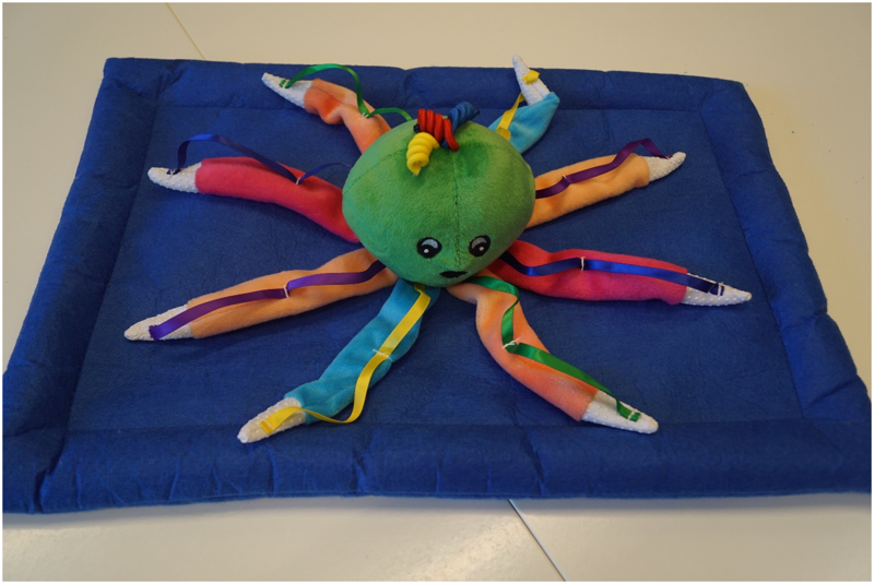 Auf einer blauen Stoffmatte ist ein Octopus (Tintenfisch) aus Stoff mit acht verschiedenfarbigen Stoffarmen befestigt. In der Mitte dieser Arme ragt ein grüner runder Kopf heraus, auf dem schwarze Augen und ein schwarzer Mund gestickt sind. Oben auf dem Kopf trägt der Octopus farbige Haarlocken. An einer Ziehschnur (die auf dem Foto nicht zu sehen ist) kann der Kopf des Octopus zum Vi­b﻿rieren gebracht werden.