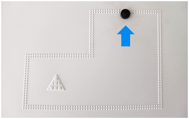 Zu sehen ist eine geometrische Figur aus zwei unterschiedlich großen Rechtecken in Form eines gespiegelten „L“, die von einer doppelten Reihe von Braillepunkten abgegrenzt ist. In der Mitte oben befindet sich eine Lücke. Durch diese Lücke kann ein Magnet hindurch geschoben werden, was durch einen blauen Pfeil in Richtung der Lücke/des Ausgangs verdeutlicht wird.