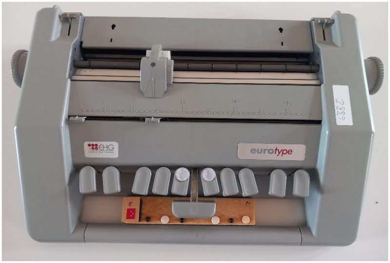 Foto einer Braillemaschine und einer Holzleiste zur Verdeutlichung der Tastenkombination, die so zwischen die untere Kante einer Eurotype-Braillemaschine geschoben wird, dass die erhabenen und nicht erhabenen Punkte im gleichen Abstand wie die Tasten angeordnet sind und somit helfen zu verdeutlichen, welche Tasten für den entsprechenden Buchstaben gedrückt werden müssen. Auf dem Foto sieht man die Holzleiste für den Buchstaben „o“, der links auf die Holzleiste aufgeklettet ist; danach folgen linear abwechselnd ein schwarzer Stecknadel- und ein weißer Reißnagelkopf, jeweils 3 an der Zahl, die in das weiche Holz hineingedrückt wurden. Diese Anordnung verdeutlicht, dass die hervorgehobenen Punkte – die durch die Stecknadelköpfe besser taktil wahrgenommen werden können – auf der darüberliegenden Tastatur gedrückt werden müssen, also die Tasten 1–3–5, um den Buchstaben „o“ ins Braillepapier zu prägen.