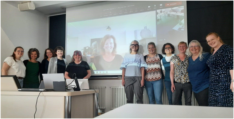 Gruppenfoto; elf Frauen stehen in einem Konferenzraum vor einer Leinwand, auf der eine weitere Frau per Videokonferenz zugeschaltet ist.