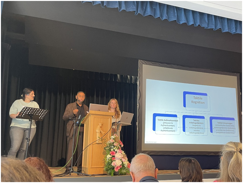 Foto mit drei Personen auf einer Bühne. Die linke Frau ist Paula Lanz und übersetzt den Vortrag von Jude Nicholas, der rechts neben ihr steht. Eine Leinwand ganz rechts zeigt die Präsentation.