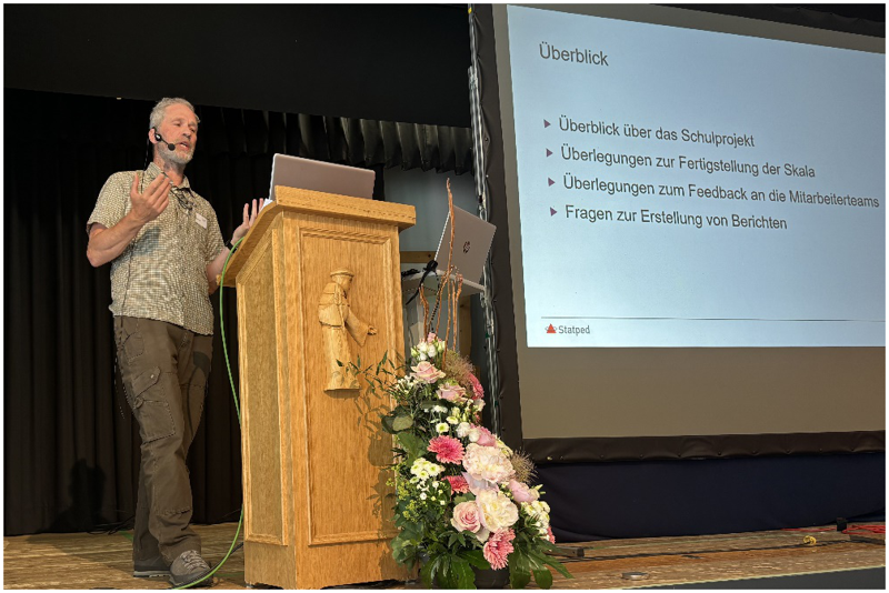 Foto von Joe Gibson auf der Bühne. Er gestikuliert, während er hinter einem Rednerpult steht. Rechts von ihm ist eine Leinwand mit der Präsentation zu sehen.