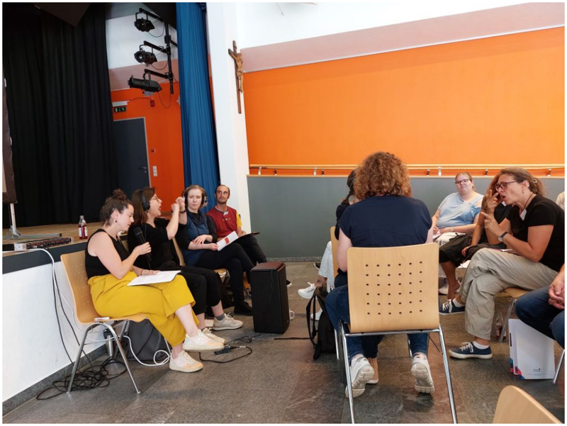 Foto von einer Kommunikationssituation. Vor der Bühne sitzen 9 Personen. Eine davon spricht in ein Mikrofon, während zwei andere Personen gebärden.