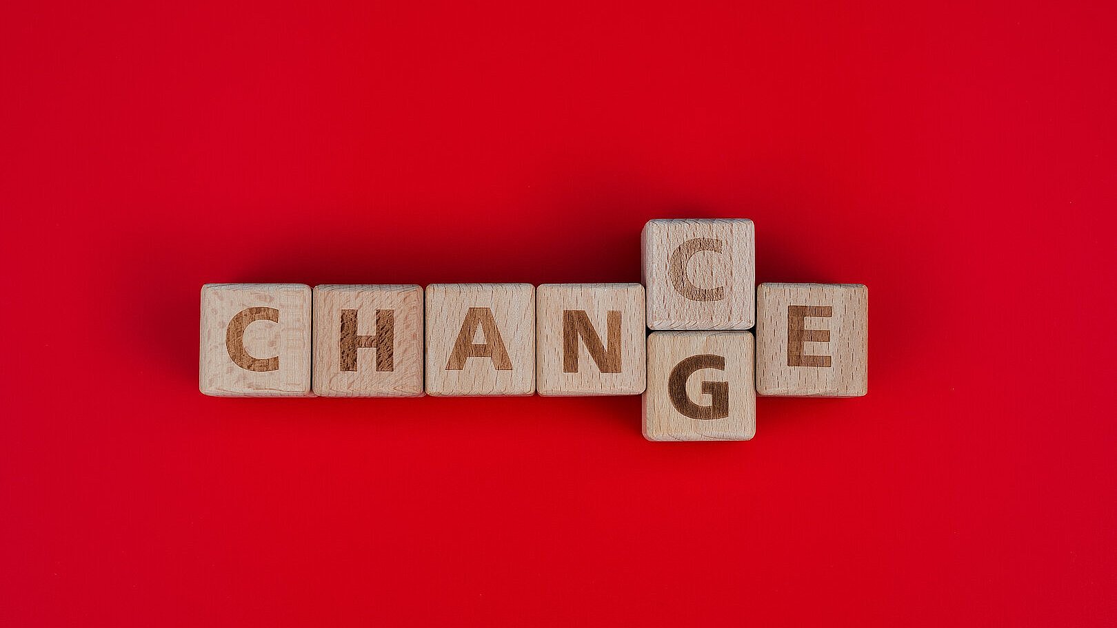 Das Bild zeigt Holzbuchstaben auf einem roten Hintergrund, die ein Kreuzworträtsel bilden: Das Wort „CHANGE“ verläuft horizontal, während das Wort „CHANCE“ vertikal dasselbe „C“ wie „CHANGE“ nutzt. Die Komposition hebt die Verbindung zwischen den Wörtern “Veränderung” und “Chance” hervor.